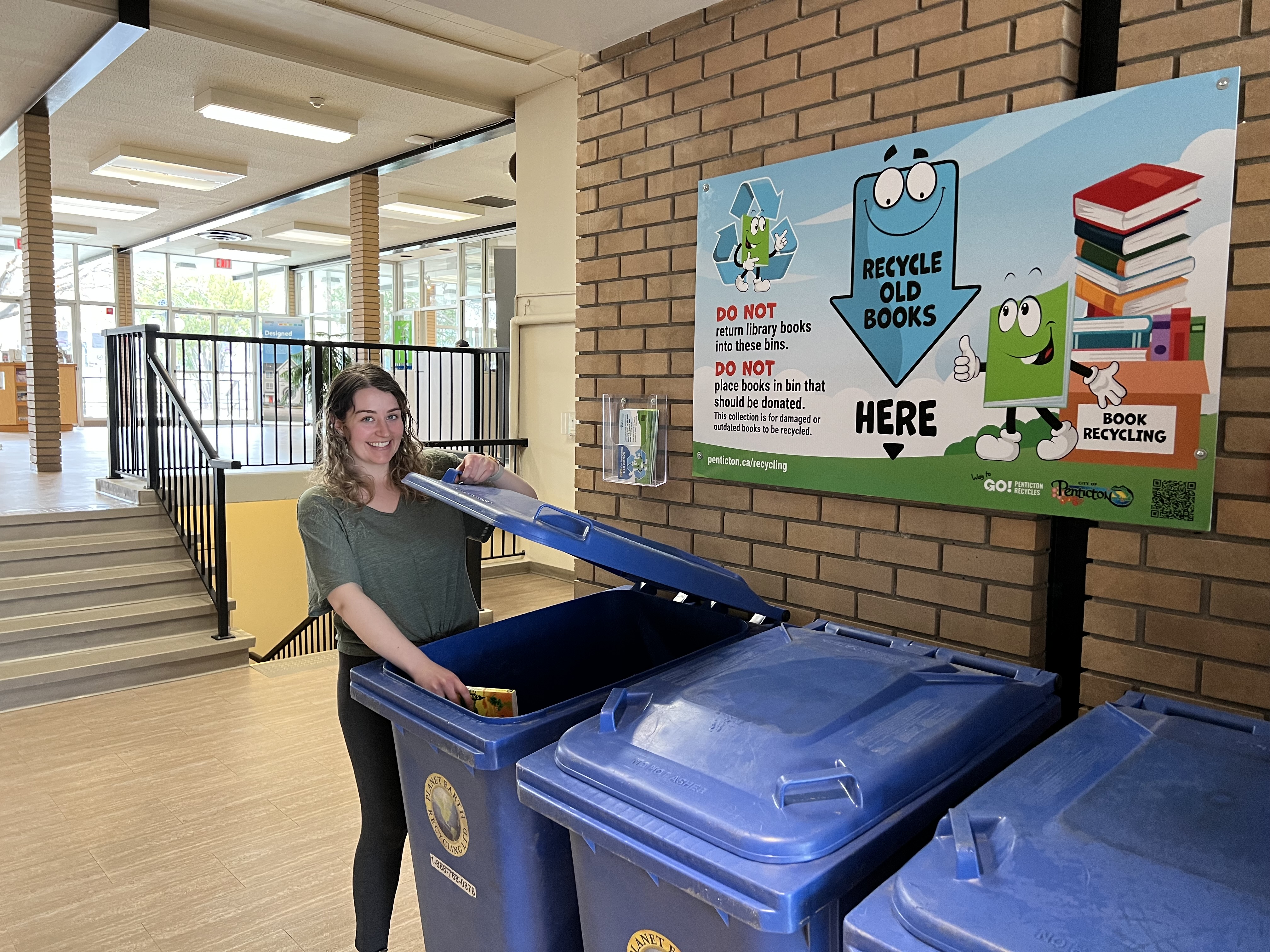 A person standing in a hallway drops a book into a blue recycling bin. A sign on the wall reads "Recycle Old Books Here," and provides guidelines for recycling. Steps lead up to a well-lit area with windows in the background.