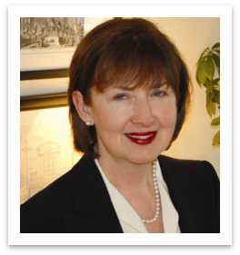 white woman with red hair in a dark suit with white blouse and pearls