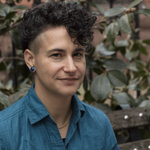 young transgender woman with dark curly hair, dark earring, blue button-up shirt, and gold chain necklace